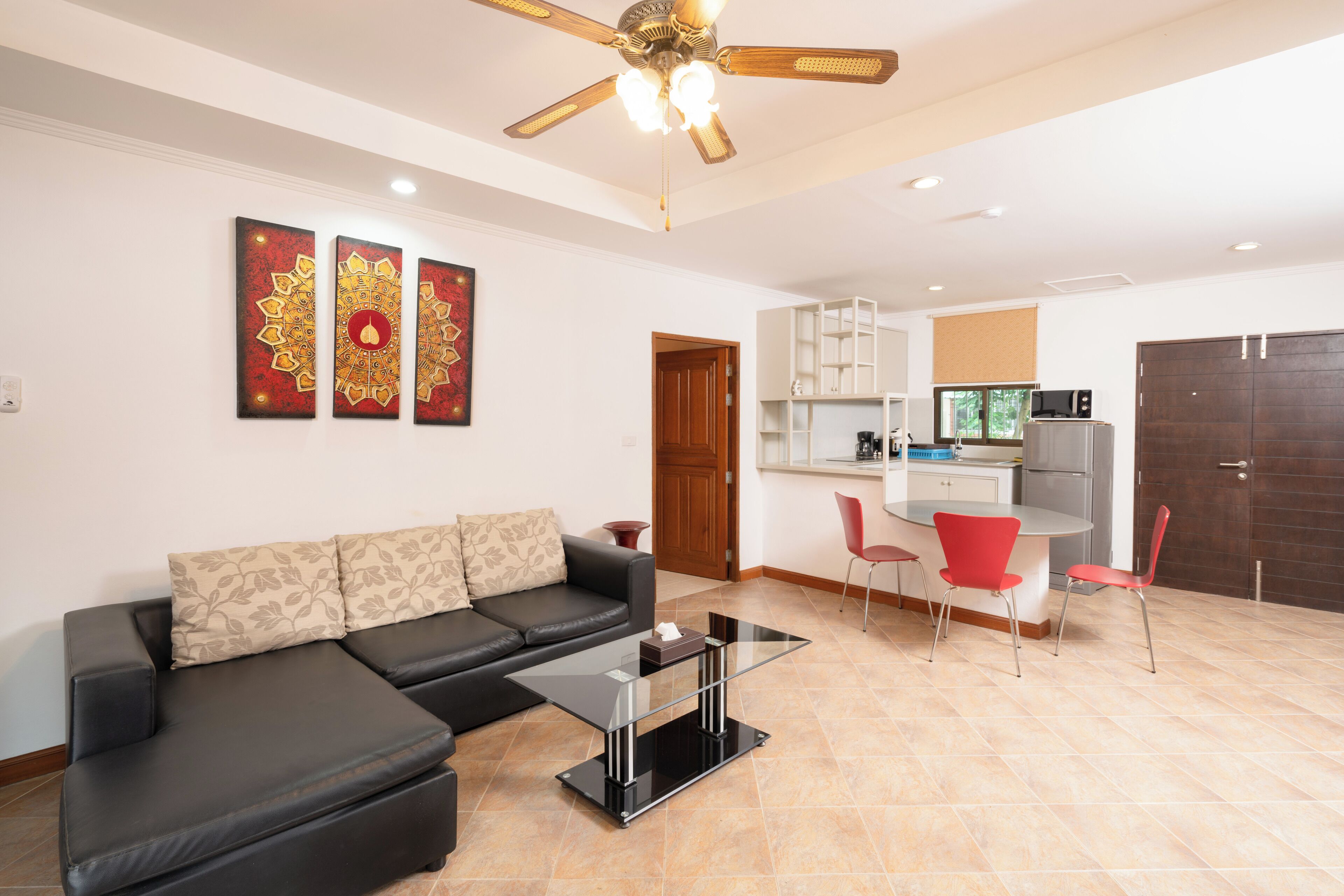 A modern living room with a black leather sofa, a glass coffee table, and a ceiling fan. There is a kitchen area with a microwave, cabinets, and a dining table with red chairs.