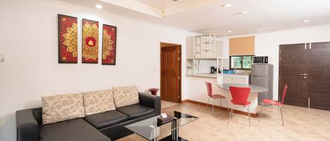 A modern living room with a black leather sofa, a glass coffee table, and a ceiling fan. There is a kitchen area with a microwave, cabinets, and a dining table with red chairs.