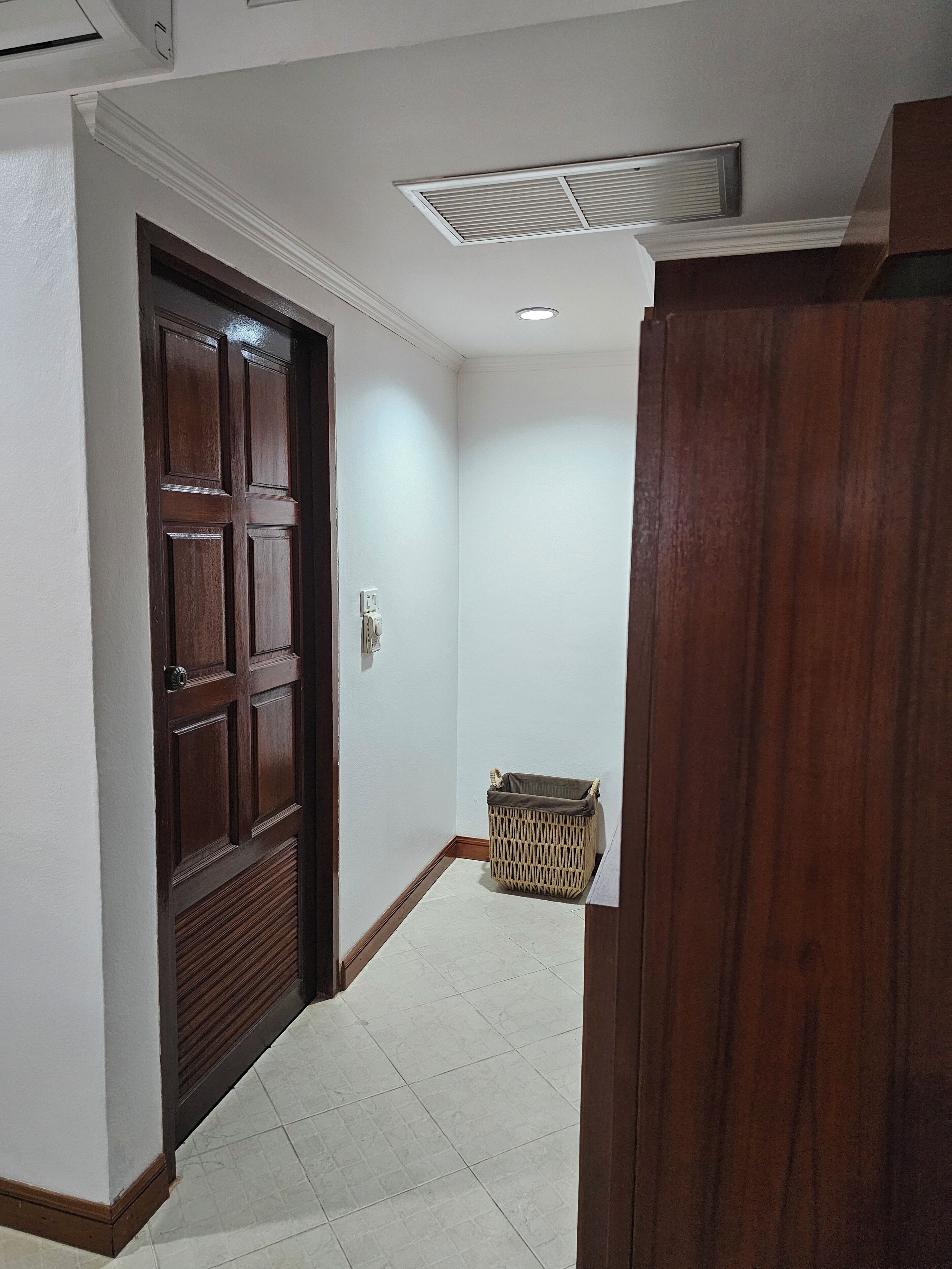 Un pasillo con una puerta de madera, un cubo de basura y una rejilla de ventilación en el techo.