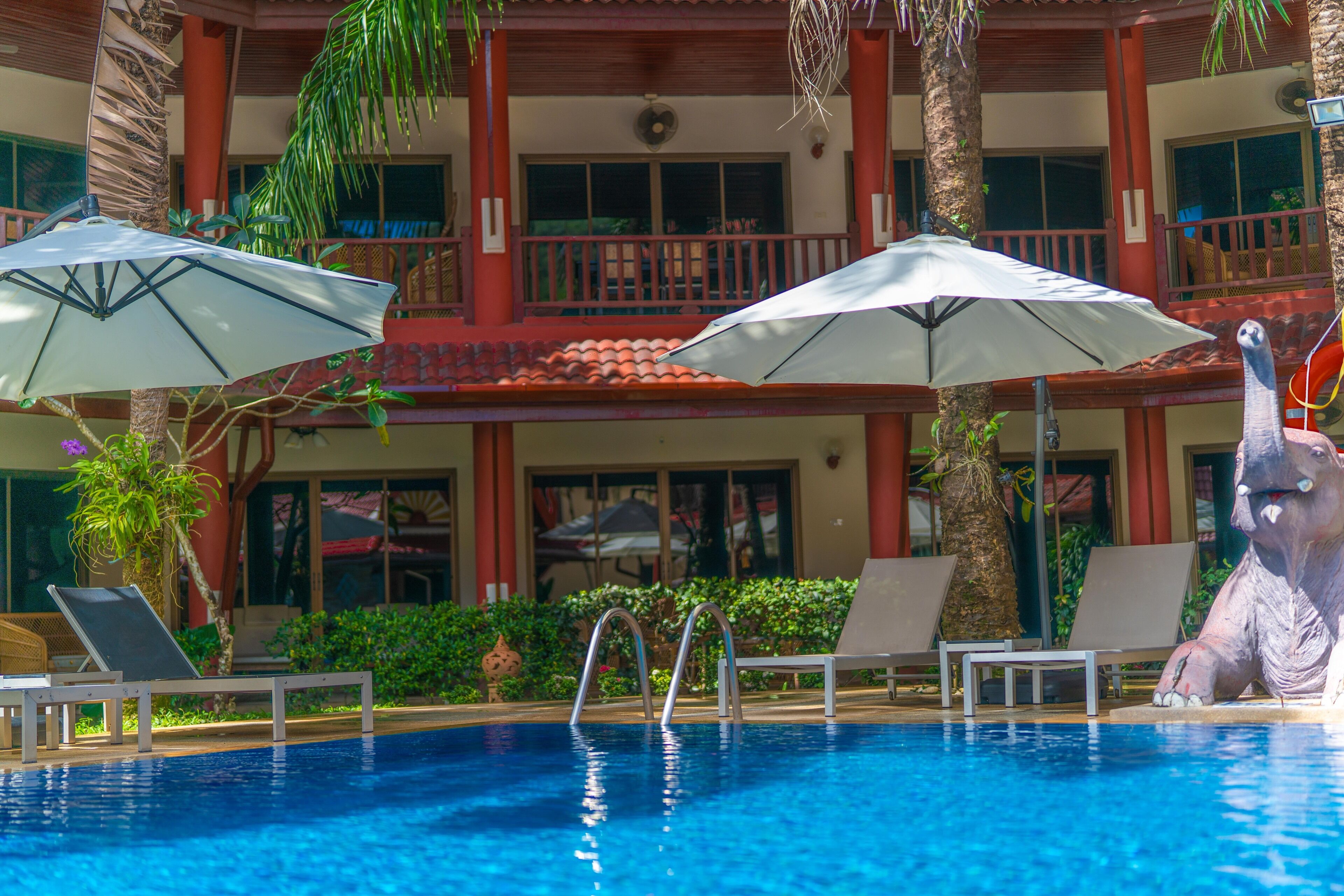 A swimming pool with lounge chairs and umbrellas, surrounded by palm trees and a building with balconies.