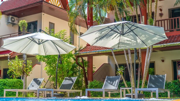 Un espace aménagé au bord de la piscine, avec des chaises longues et des parasols, entouré de palmiers et bordé d’un bâtiment dont le toit est recouvert de tuiles rouges.