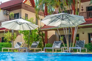 A poolside area with lounge chairs and umbrellas, surrounded by palm trees and a building with a red-tiled roof. - Sai Rougn Apartment (Patong)
