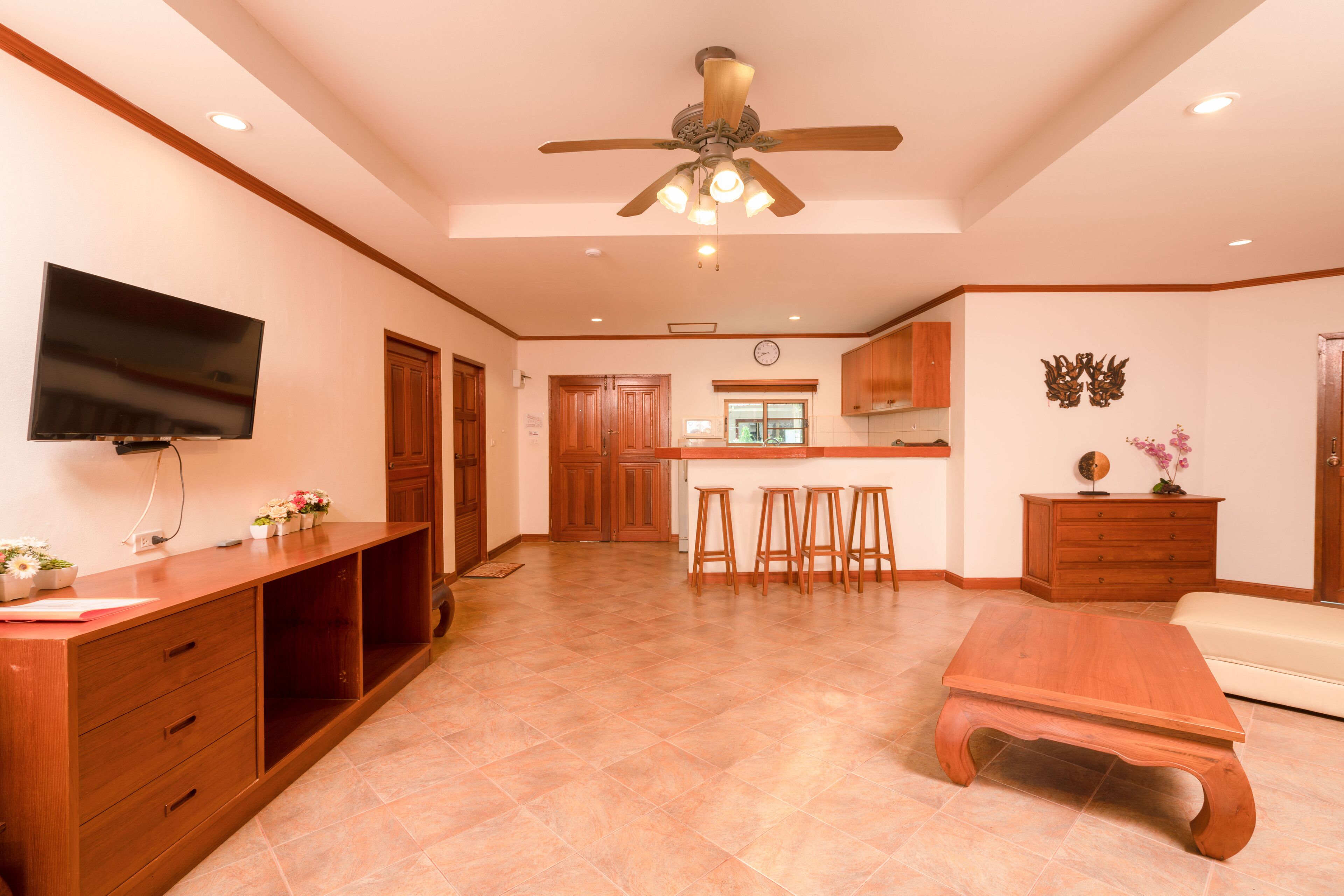 A spacious living area with a wooden entertainment unit, a flat-screen TV, a ceiling fan, and a kitchen in the background.
