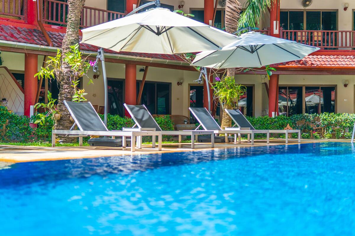 A pool area with lounge chairs and umbrellas, surrounded by a building with a red roof and columns.