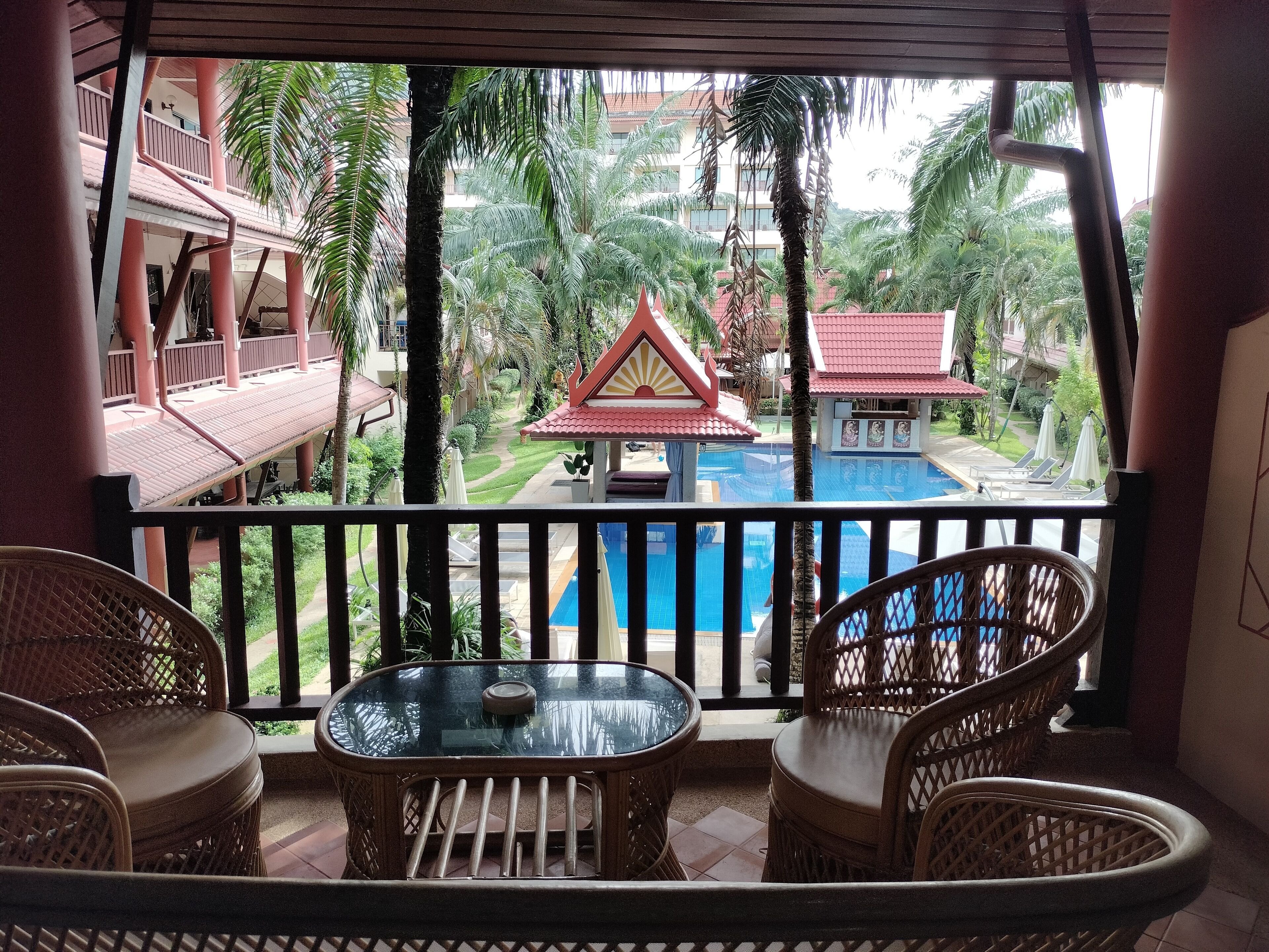 A balcony with wicker furniture overlooking a pool and a traditional building.