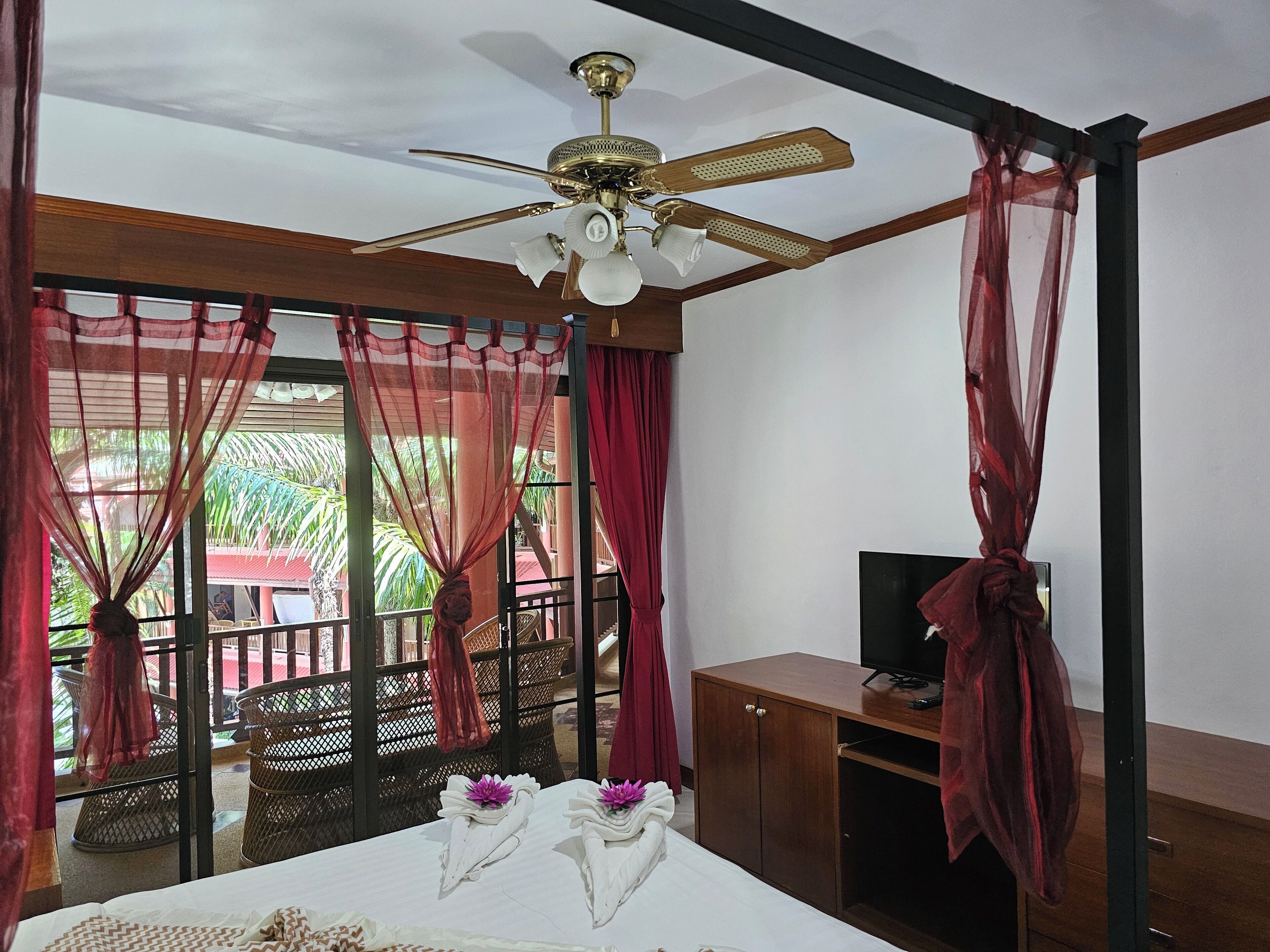 Habitación de hotel con cama, ventilador de techo, televisor y vista al balcón exterior.