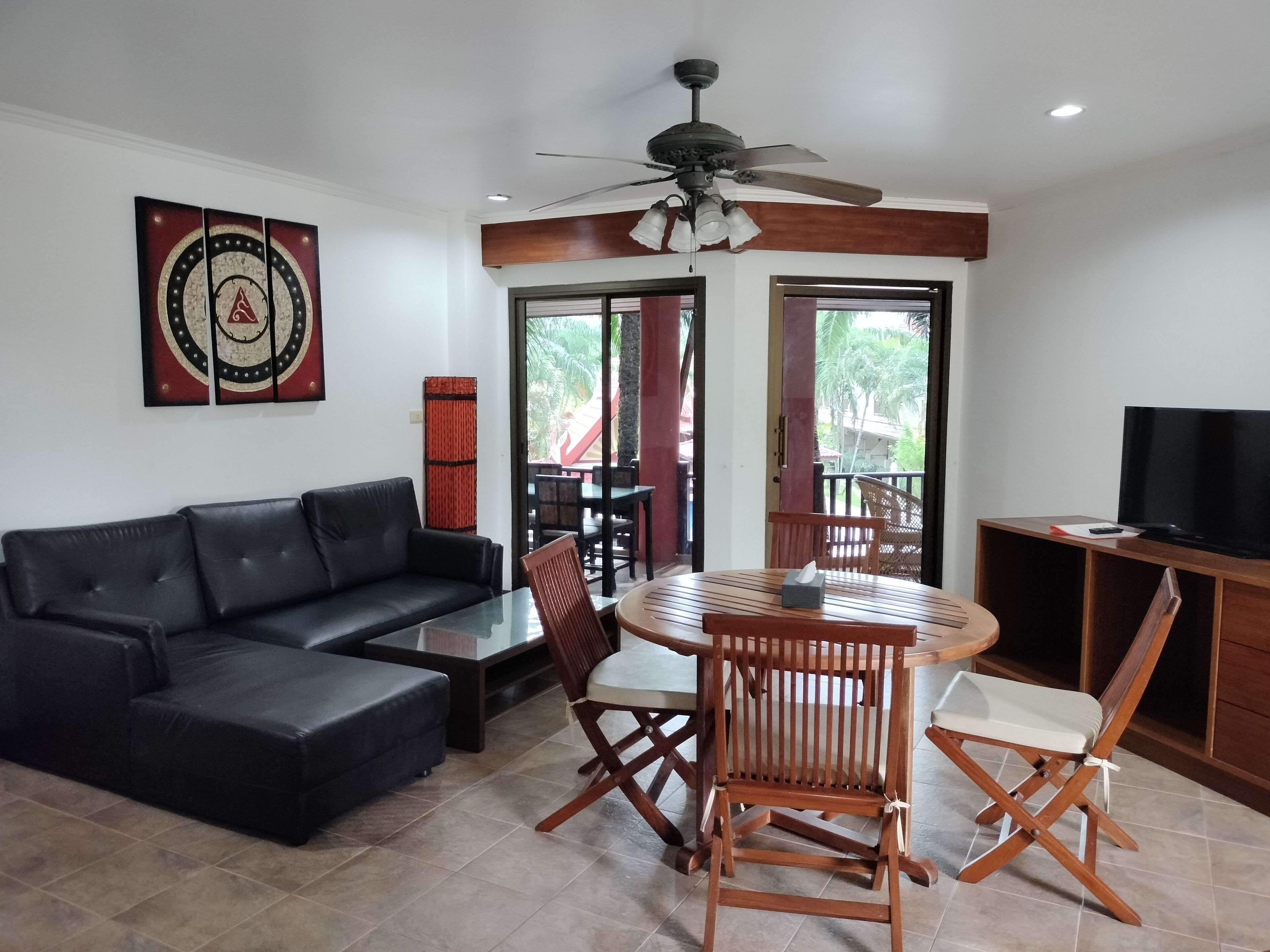 A living room with a black leather sofa, a glass coffee table, wooden chairs, a ceiling fan, and a television on a wooden stand.