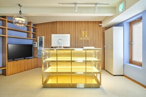 A modern hotel reception area with a wooden panel wall, a reception desk with a glass display, a flat-screen TV, and a digital kiosk.