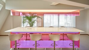 A modern dining area with a long table and four chairs, a potted plant, and a window with sheer curtains.