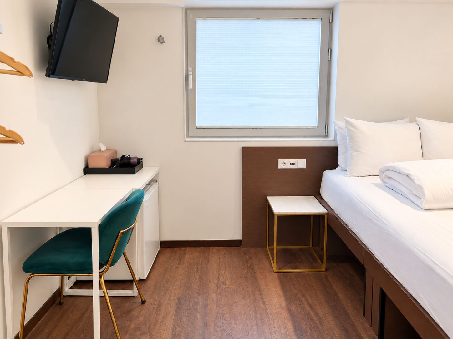 A compact hotel room with a bed, a desk, a chair, a television, and a window with blinds.