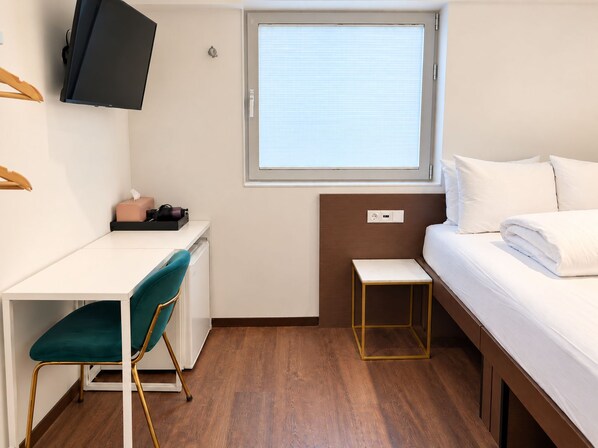 A compact hotel room with a bed, a desk, a chair, a television, and a window with blinds.