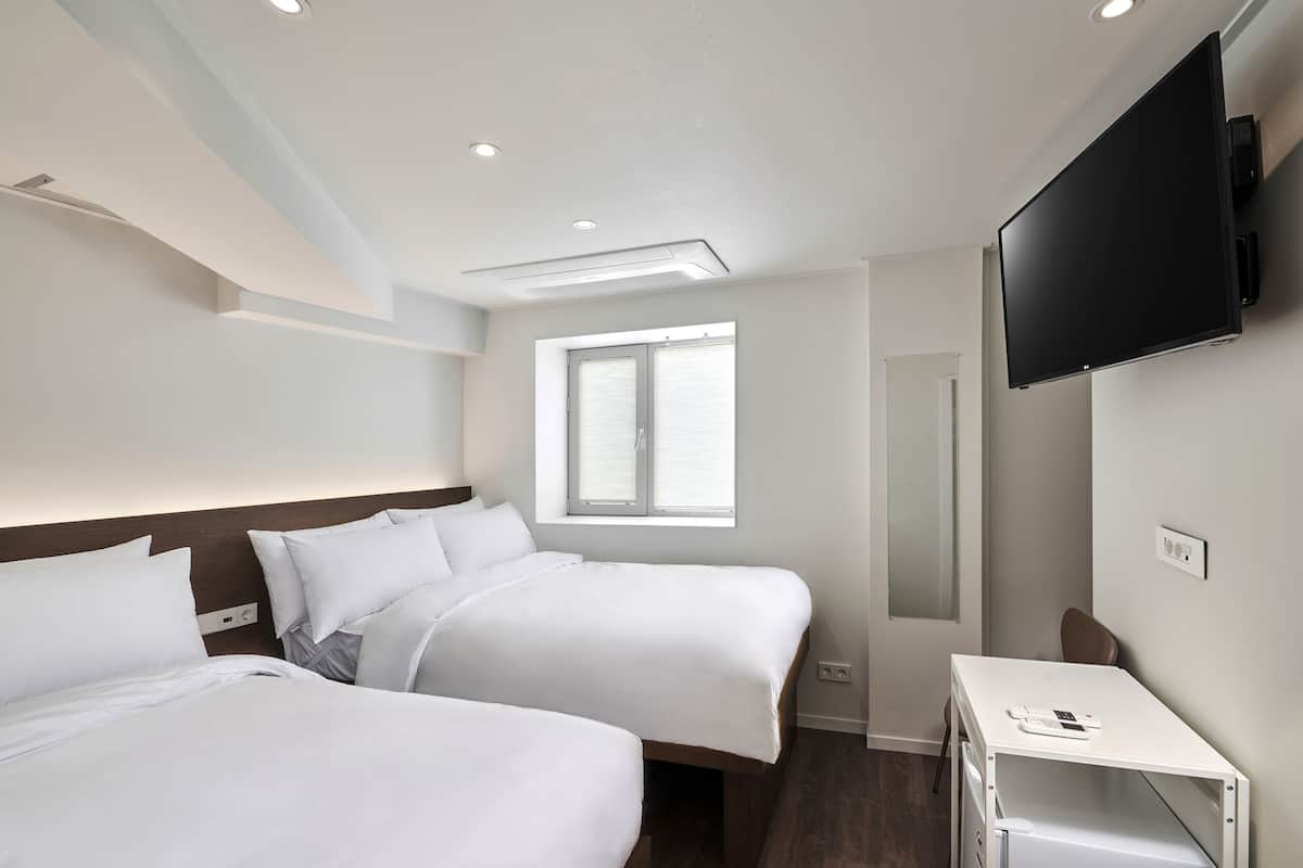 Habitación de hotel con dos camas, un televisor montado en la pared, una ventana y un escritorio pequeño con un teléfono.