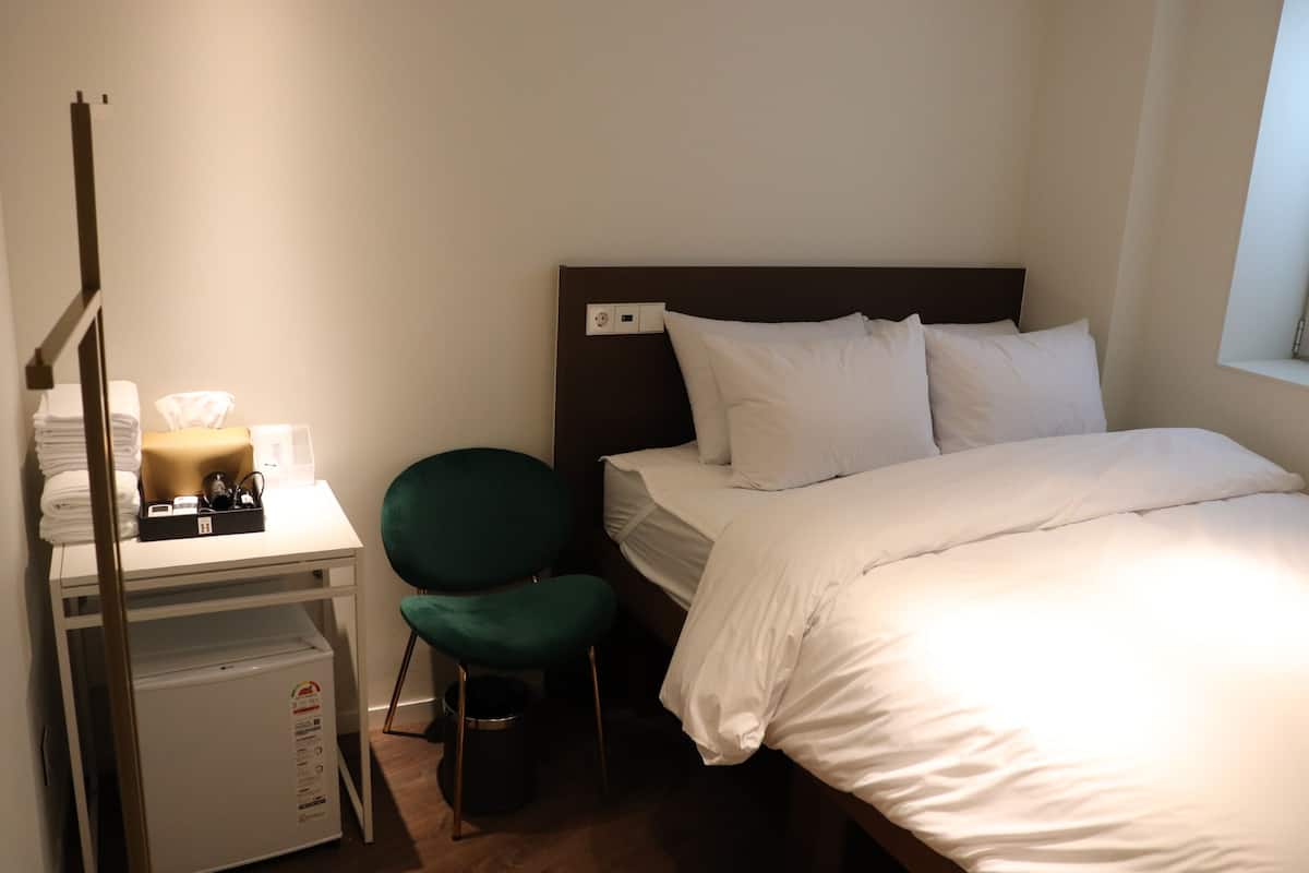 Habitación de hotel con cama, escritorio y silla, una mesita de noche con lámpara, y un enchufe.