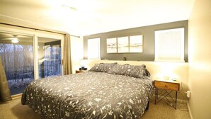 Een slaapkamer met een groot bed, een nachtkastje, een lamp, een raam met jaloezieën en uitzicht op bomen.