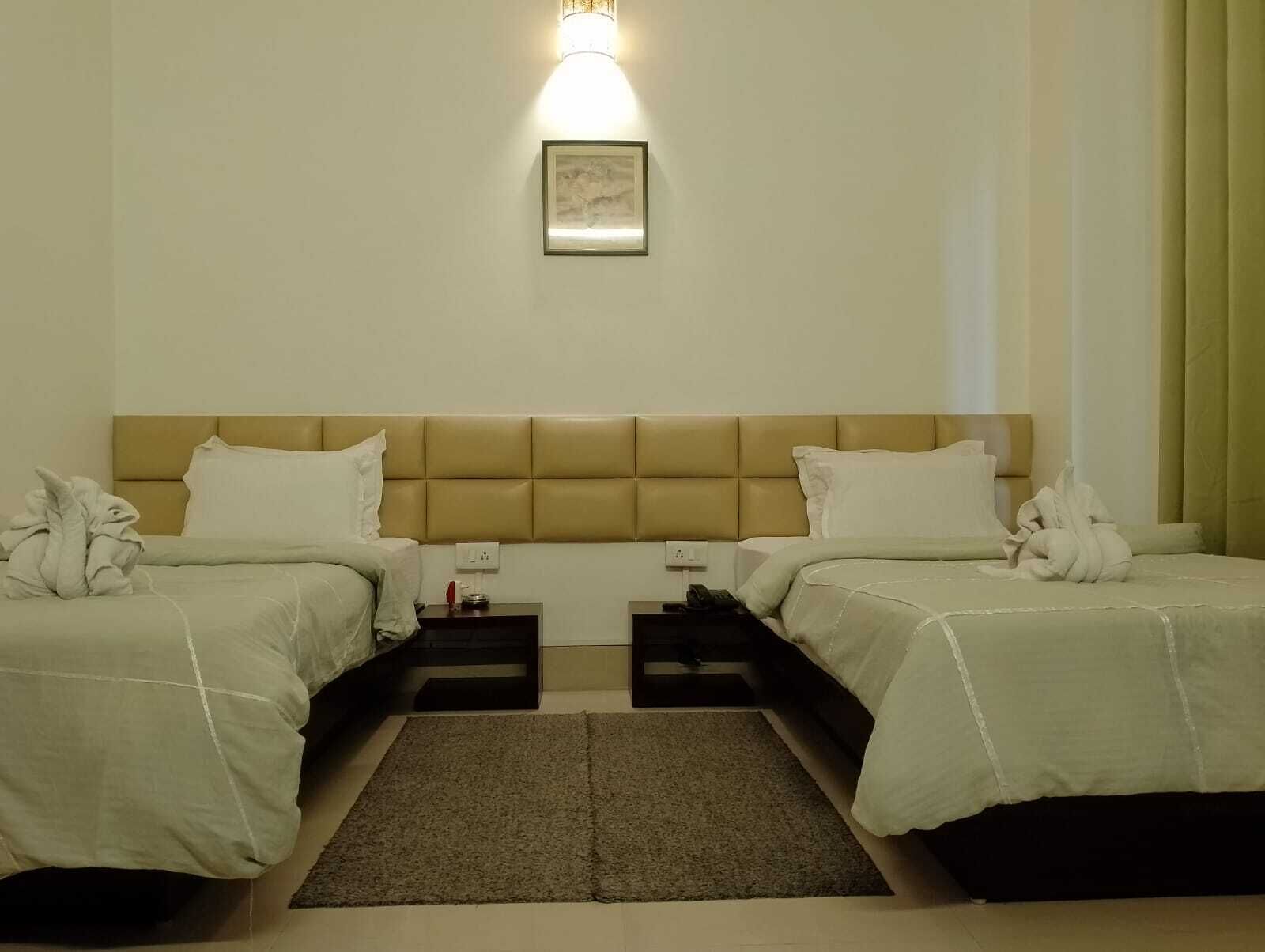 A hotel room with two beds, a headboard, a bedside table with a lamp, a framed picture on the wall, and a green curtain.