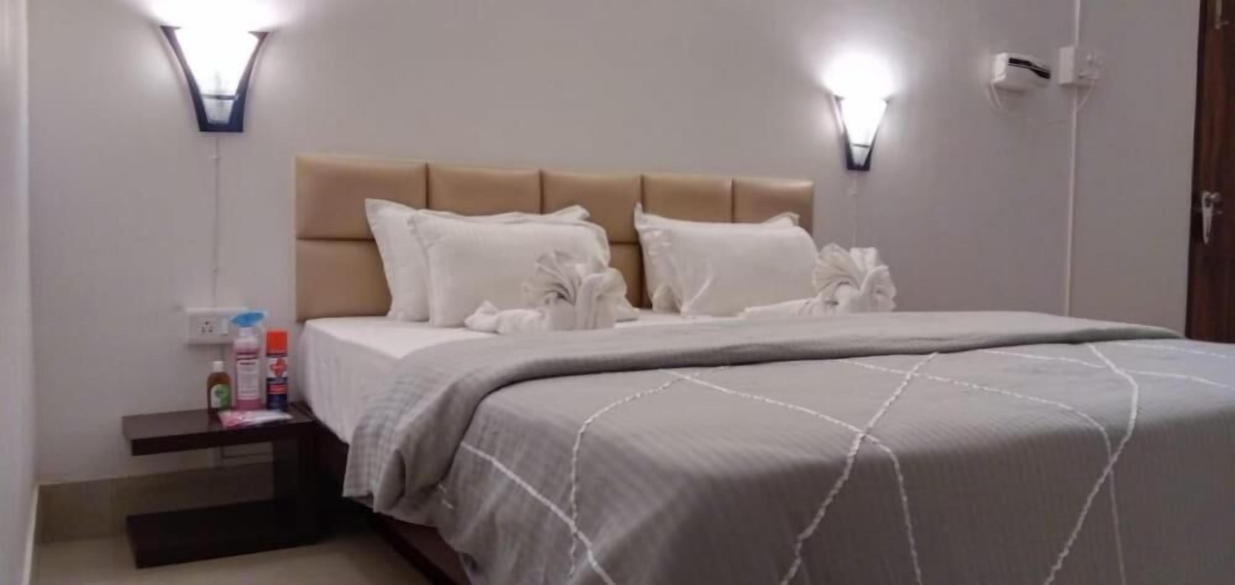 A neatly made bed with a beige headboard, white pillows, and a grey bedspread. There is a nightstand with toiletries and a wall-mounted light fixture.