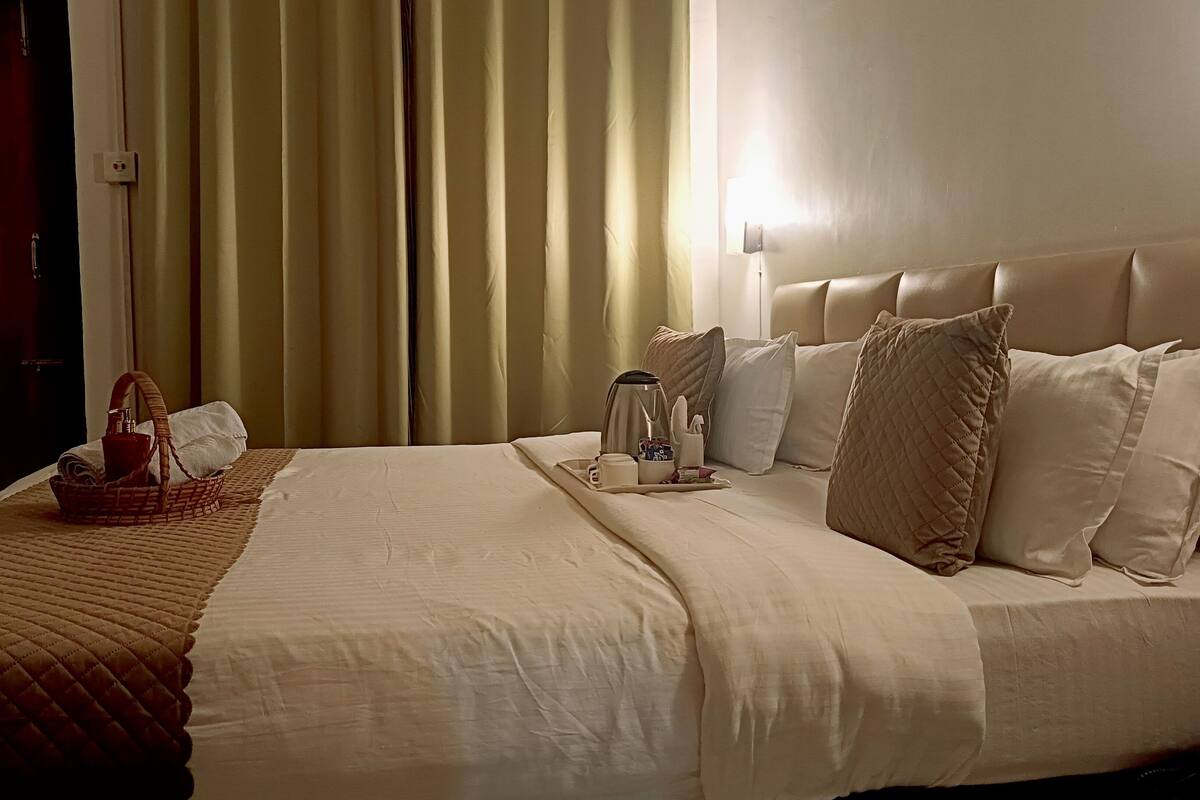 A neatly made bed with a quilted cover, a bedside table with a lamp, and a basket with a bottle and a tissue box.