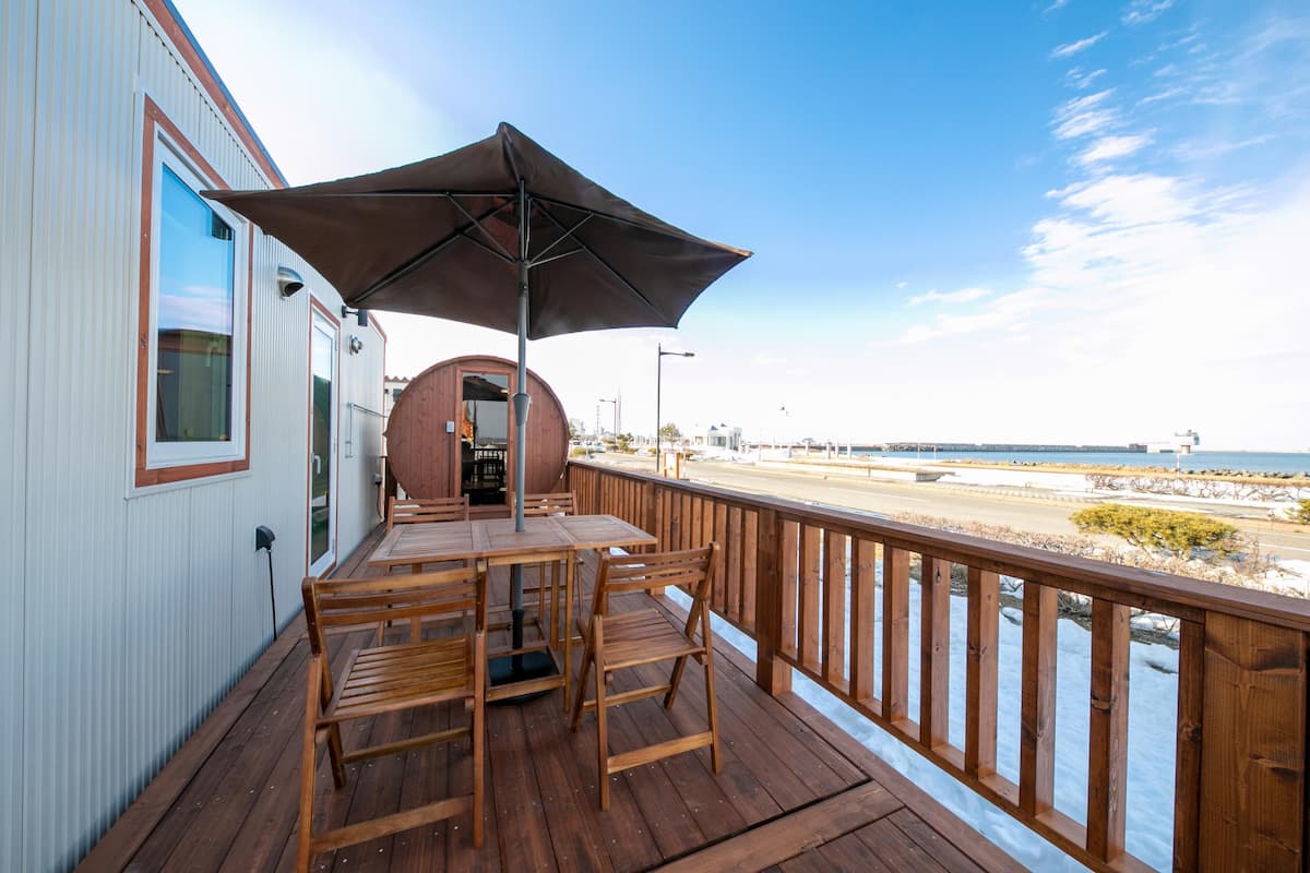 A wooden deck with tables and chairs, a large umbrella, and a view of the beach and ocean.