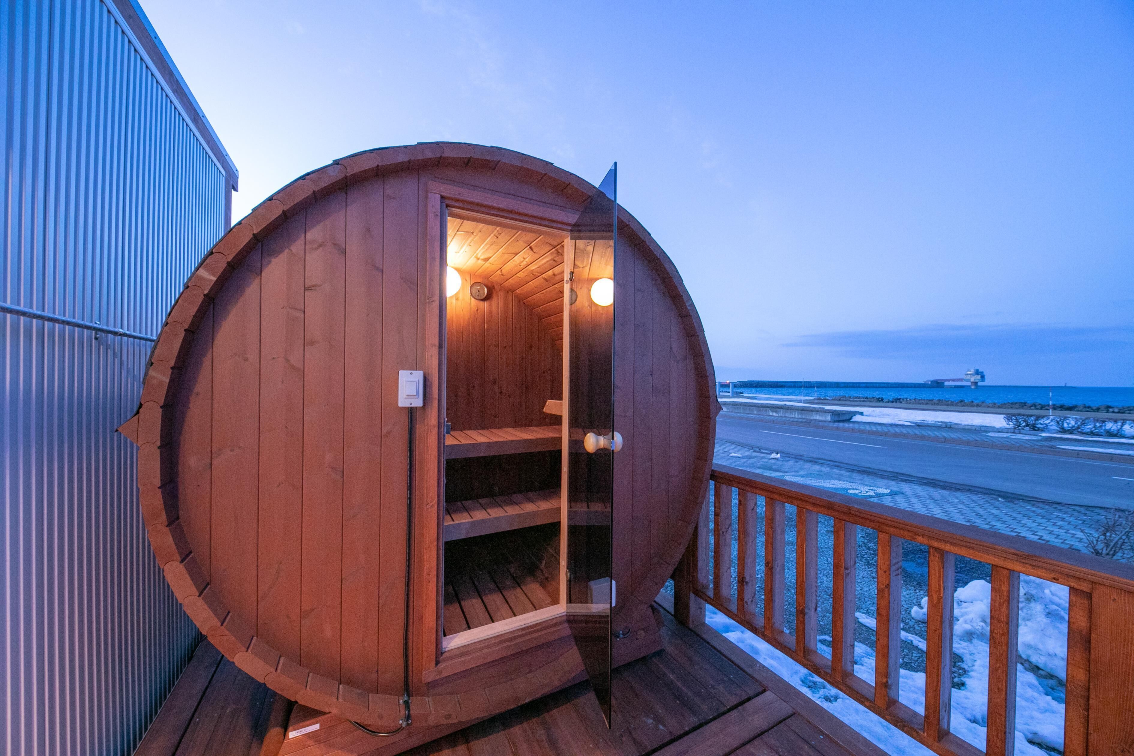 A wooden sauna structure with a glass door, a wooden deck, and a view of the beach.