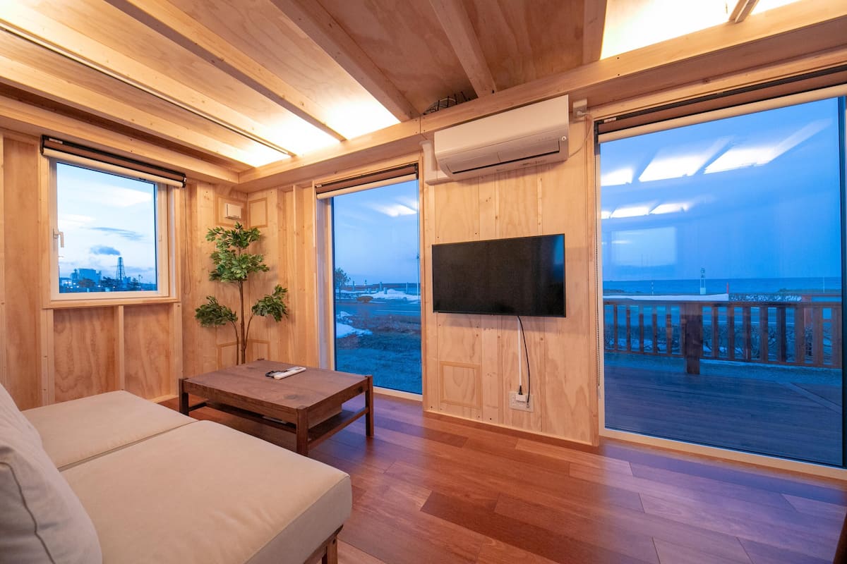 A wooden interior with a sofa, a table, a TV, and large windows overlooking a snowy landscape.