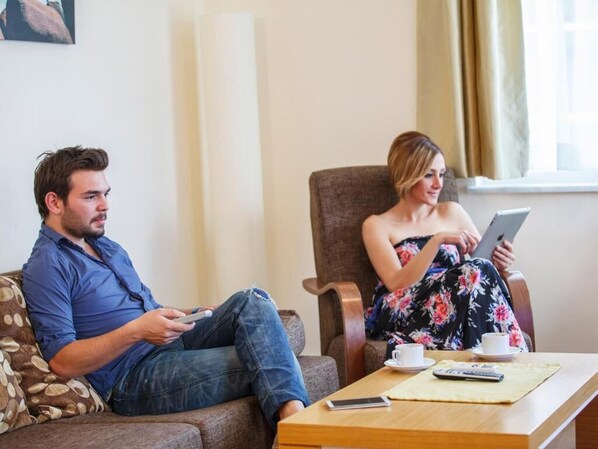 Un homme et une femme sont assis sur un canapé et une chaise dans une pièce avec une fenêtre, une table sur laquelle se trouvent une tasse et une télécommande.