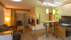 A modern hotel room with a sofa, a small table, a TV, a kitchenette, and a dining area.