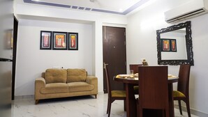 A living room with a sofa, dining table, chairs, and framed pictures on the wall. - Lime Tree 2Bhk Serviced Apartment (Gurugram)