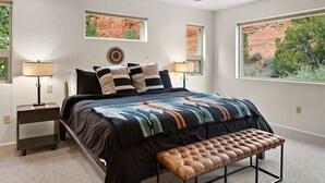 A bedroom with a bed, two bedside lamps, a bench, and windows with a view of red rock and greenery.