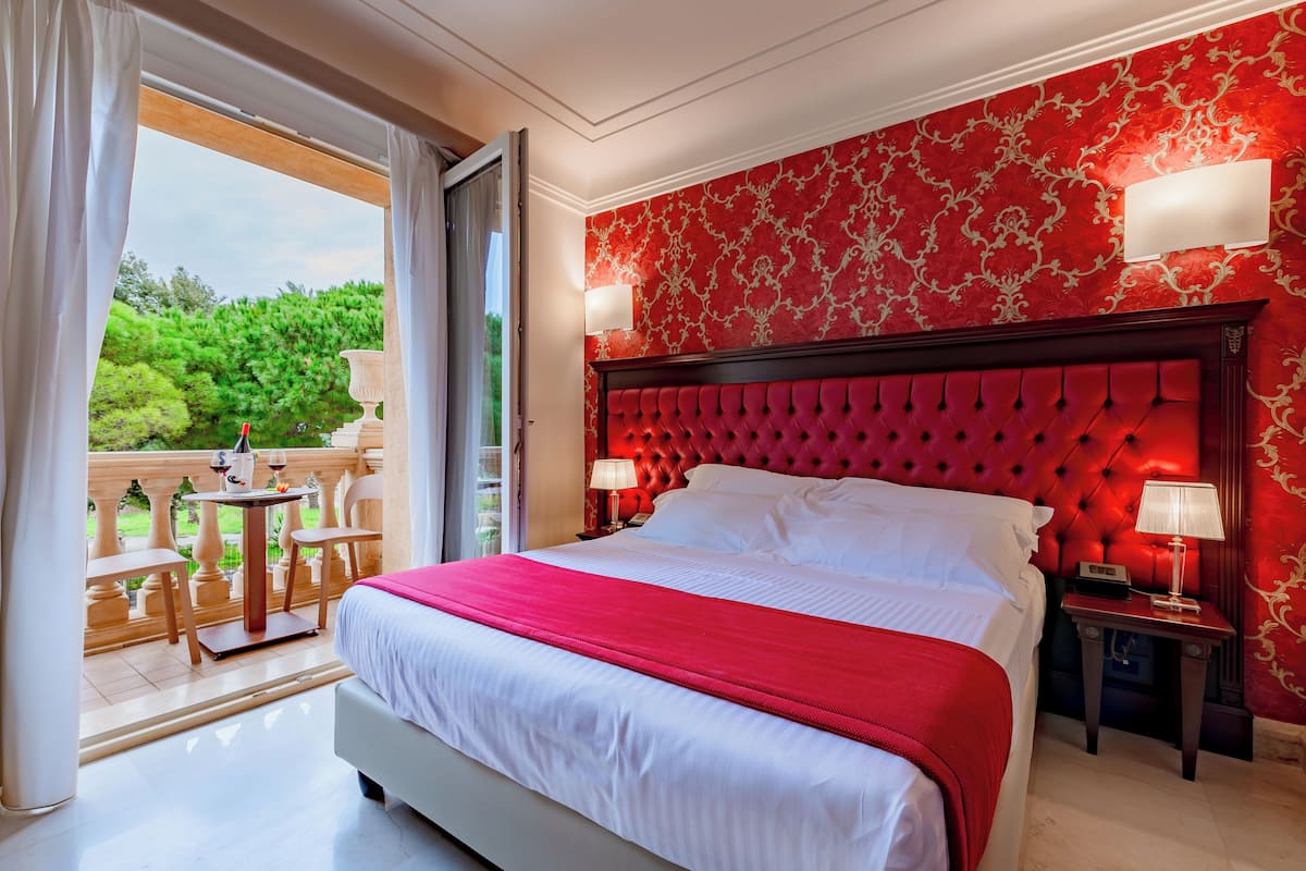 A hotel room with a large bed, a balcony with a table and chairs, and a red patterned wall.
