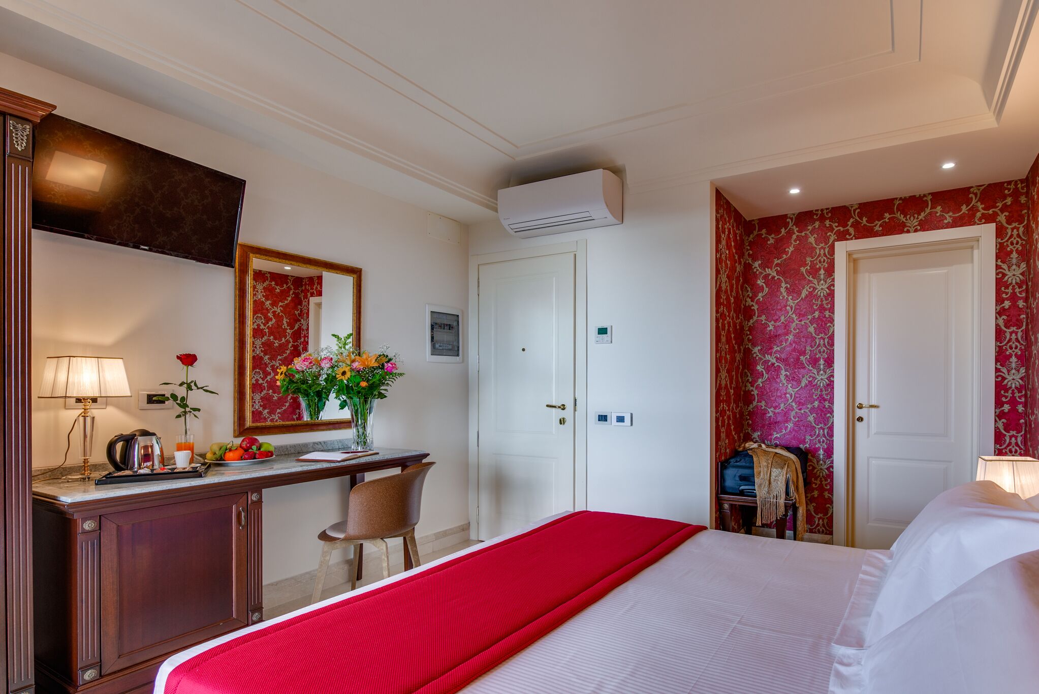 A hotel room with a bed, a desk with a lamp, a mirror, a dresser, and a wardrobe.