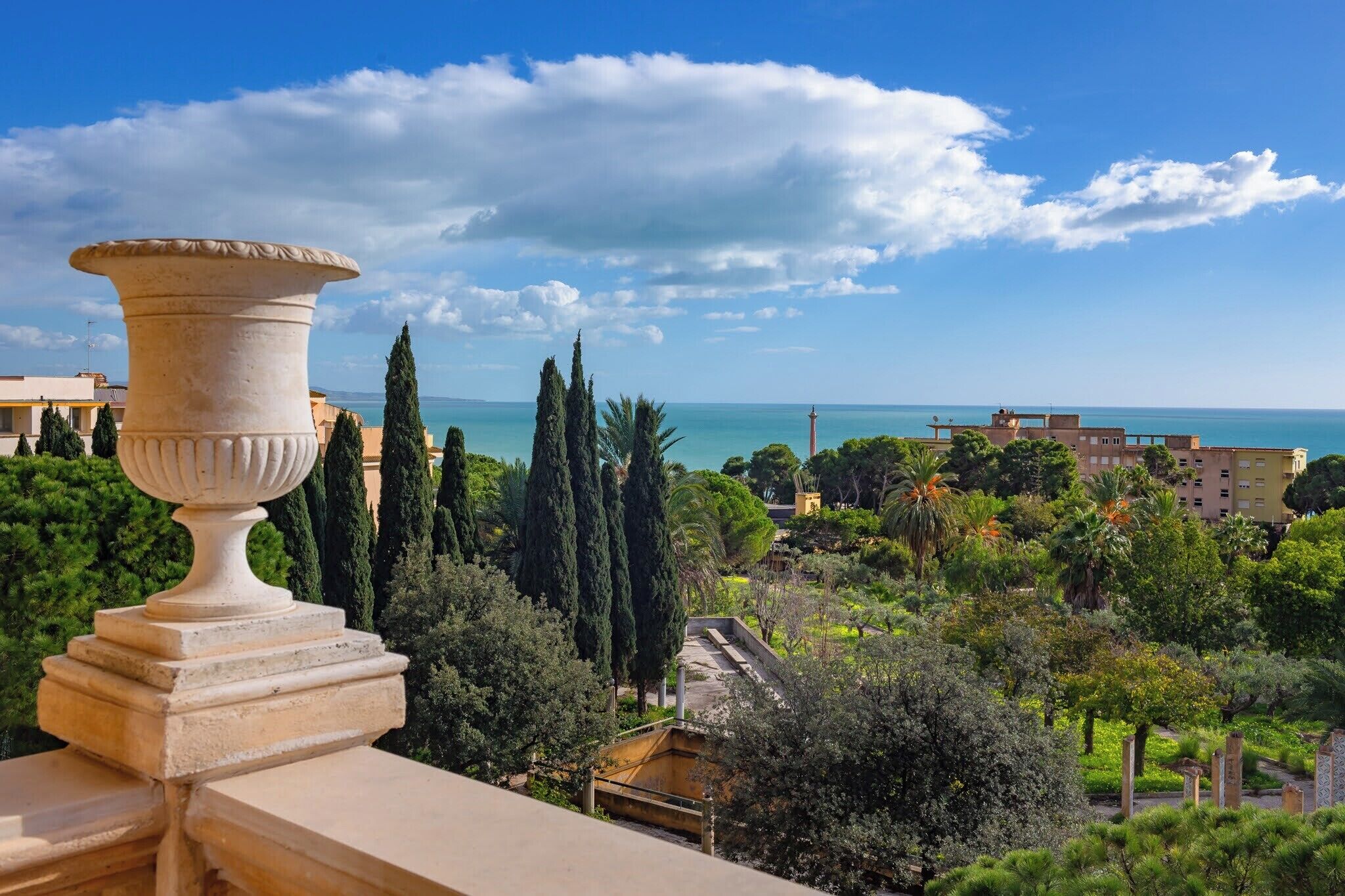 Vista di una zona costiera con un'ampia urna in pietra su un balcone, vegetazione rigogliosa e un edificio in lontananza con vista sul mare.