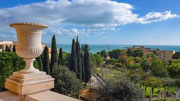 Vista di una zona costiera con un'ampia urna in pietra su un balcone, vegetazione rigogliosa e un edificio in lontananza con vista sul mare.