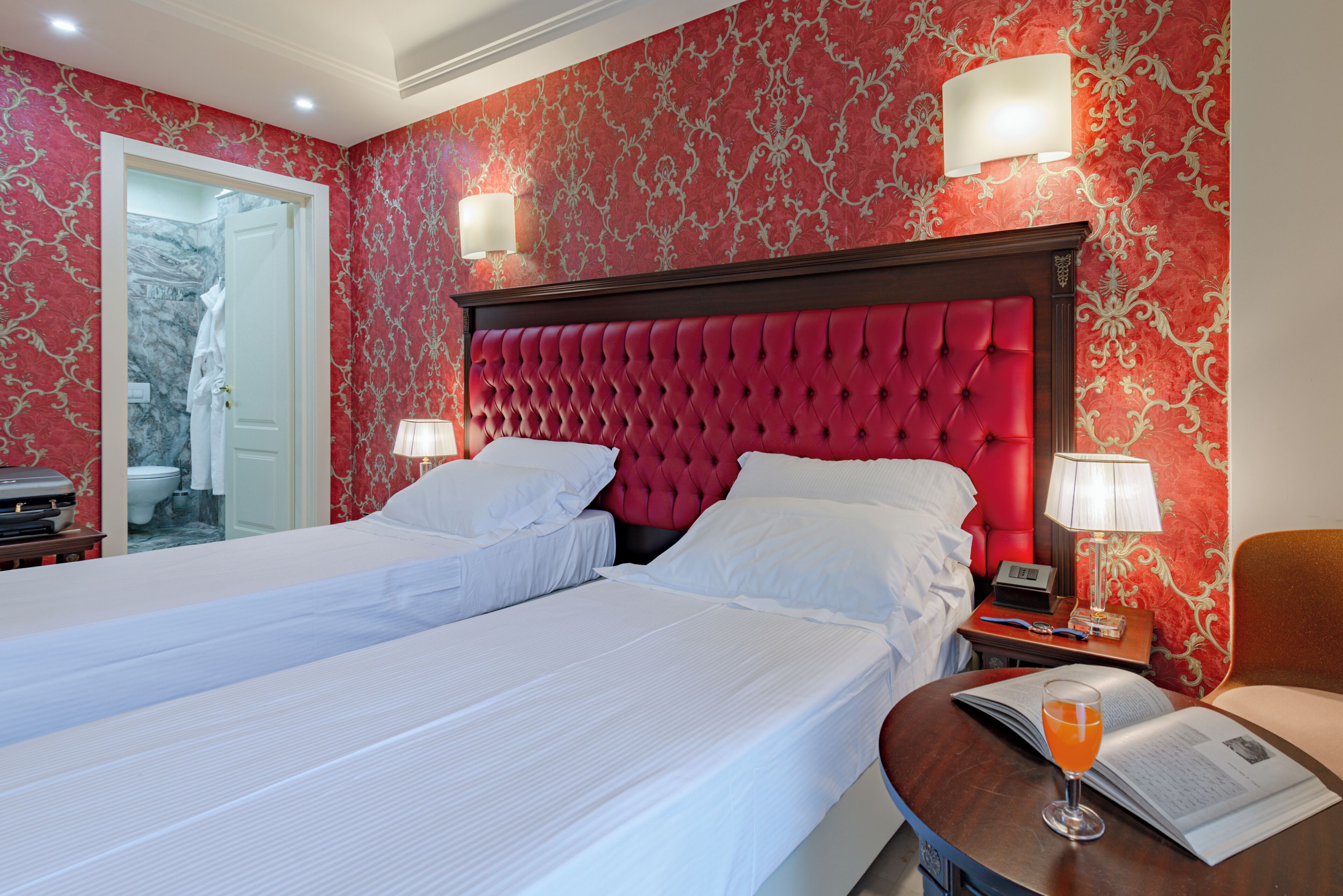 A hotel room with two beds, a red headboard, a bedside table with a lamp, and a glass of orange juice on a small table.