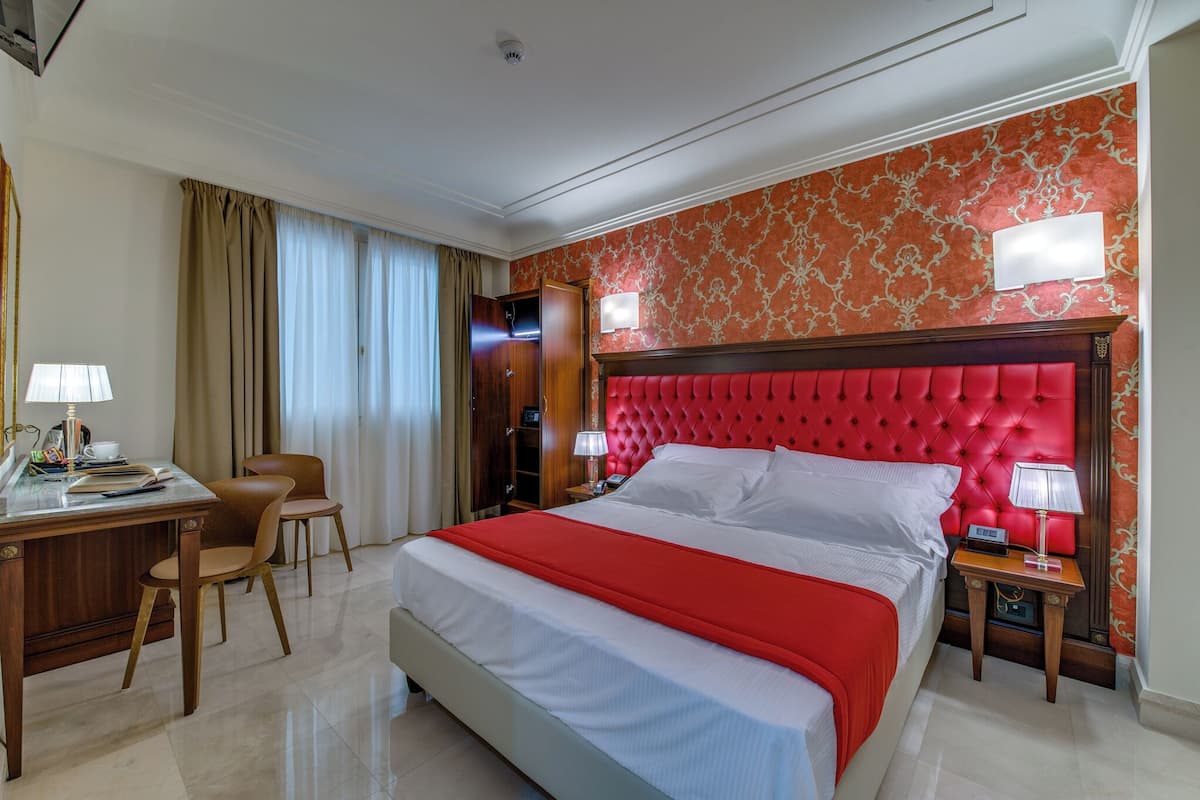 A hotel room with a large bed, a desk with a chair, a wardrobe, and a nightstand with a lamp.