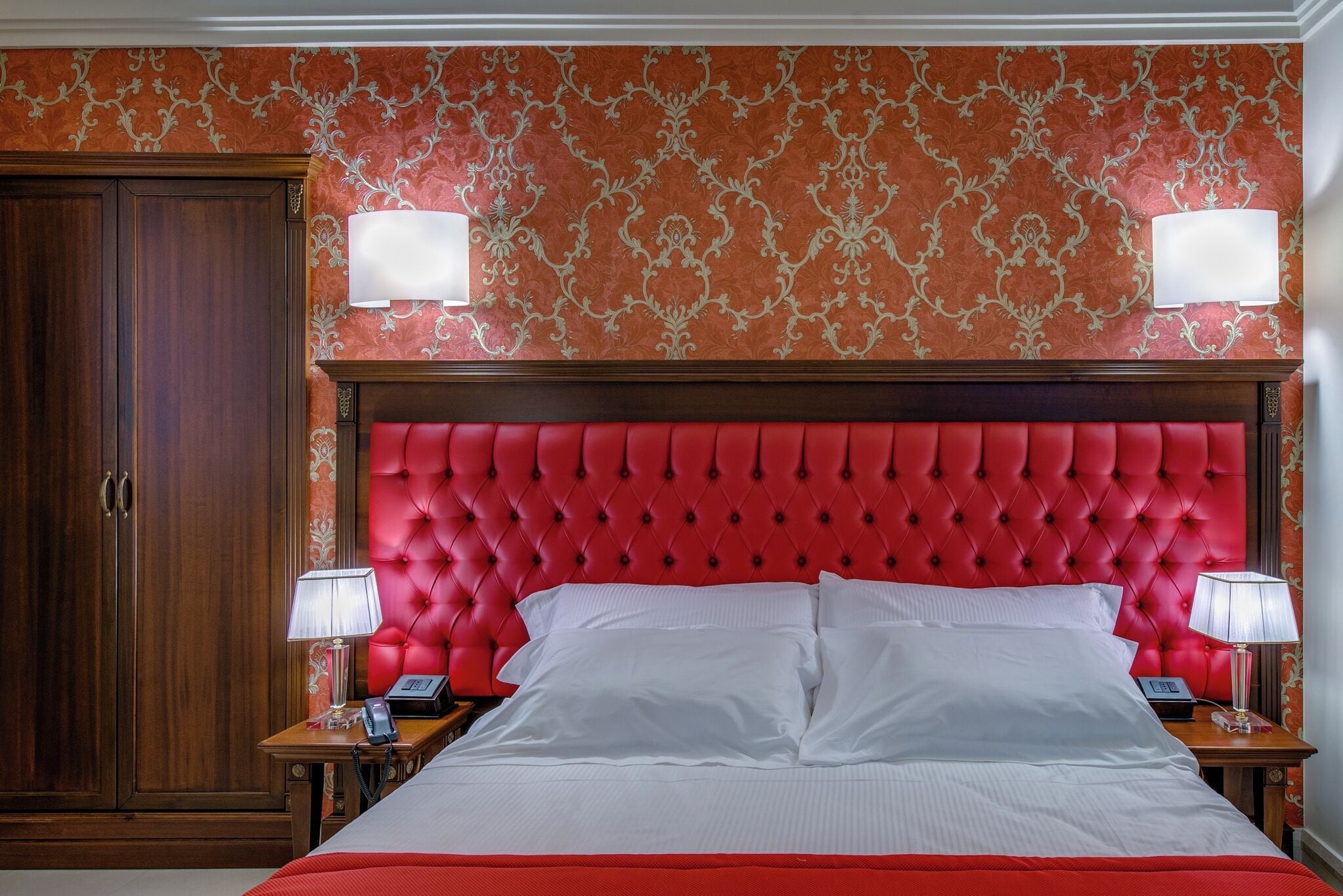 A hotel room with a red tufted headboard, a large bed with white linens, two bedside tables with lamps, and a wooden wardrobe.