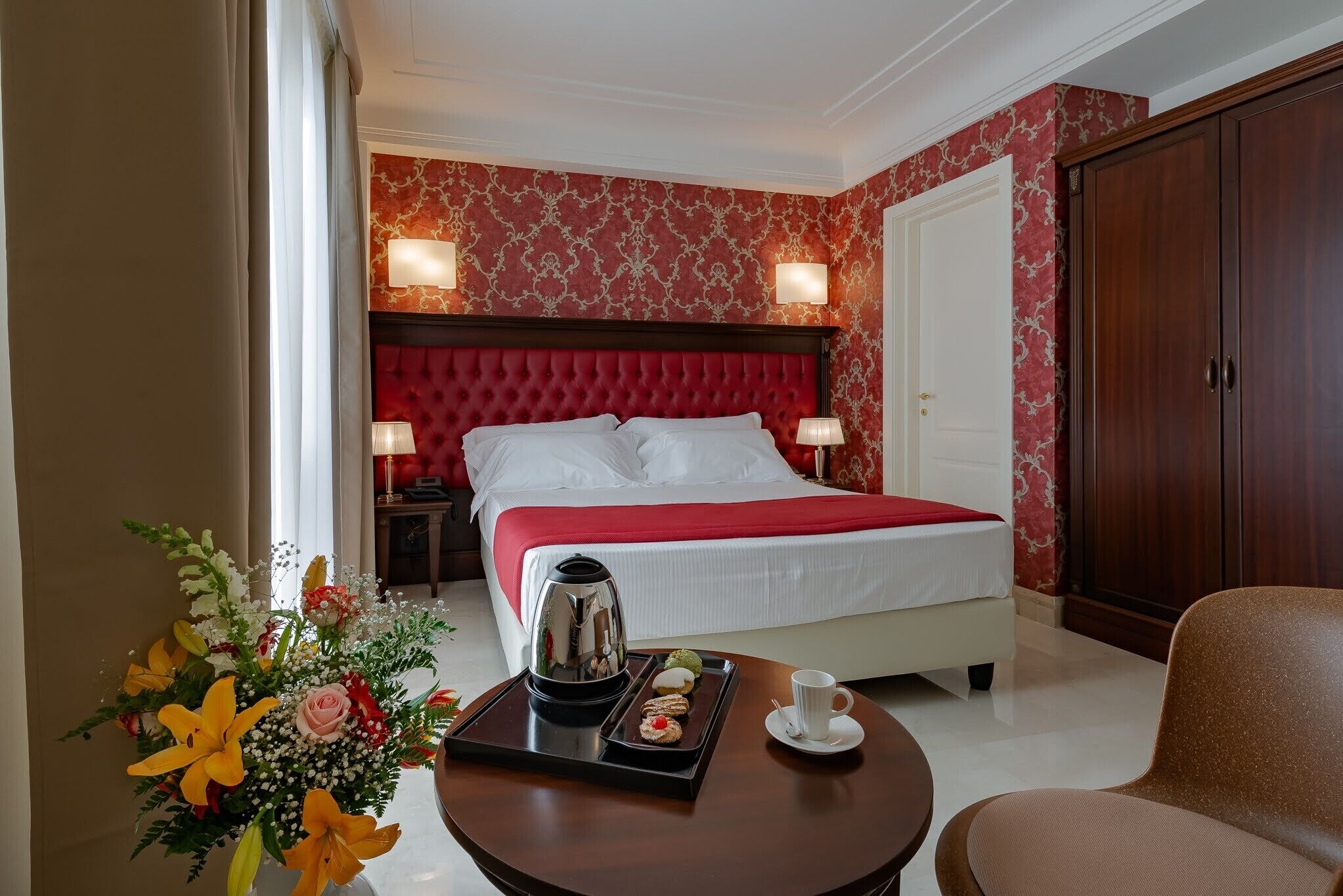 A hotel room with a red upholstered headboard, a bed with white linens, a bedside table with a tray of snacks, a coffee pot, and a floral arrangement.
