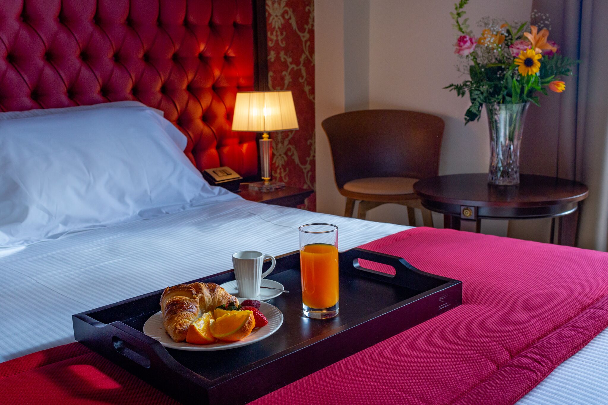 A hotel room with a bed, a bedside table with a lamp, a chair, a small table with a vase of flowers, and a tray with breakfast items including croissants, fruit, and orange juice.