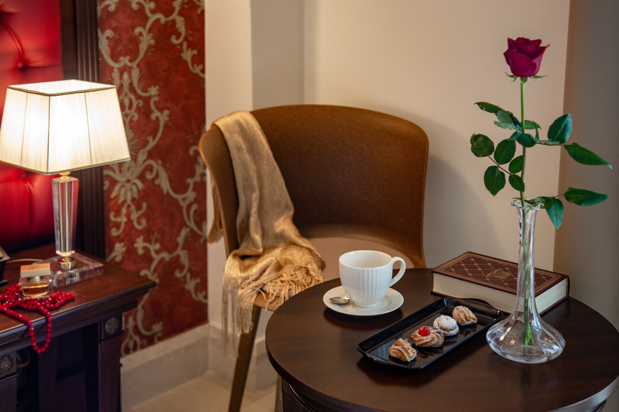 A cozy room with a plush red chair, a white cup on a saucer, a tray of pastries, a vase with a rose, and a lamp on a side table.