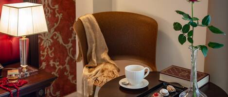 A cozy room with a plush red chair, a white cup on a saucer, a tray of pastries, a vase with a rose, and a lamp on a side table.
