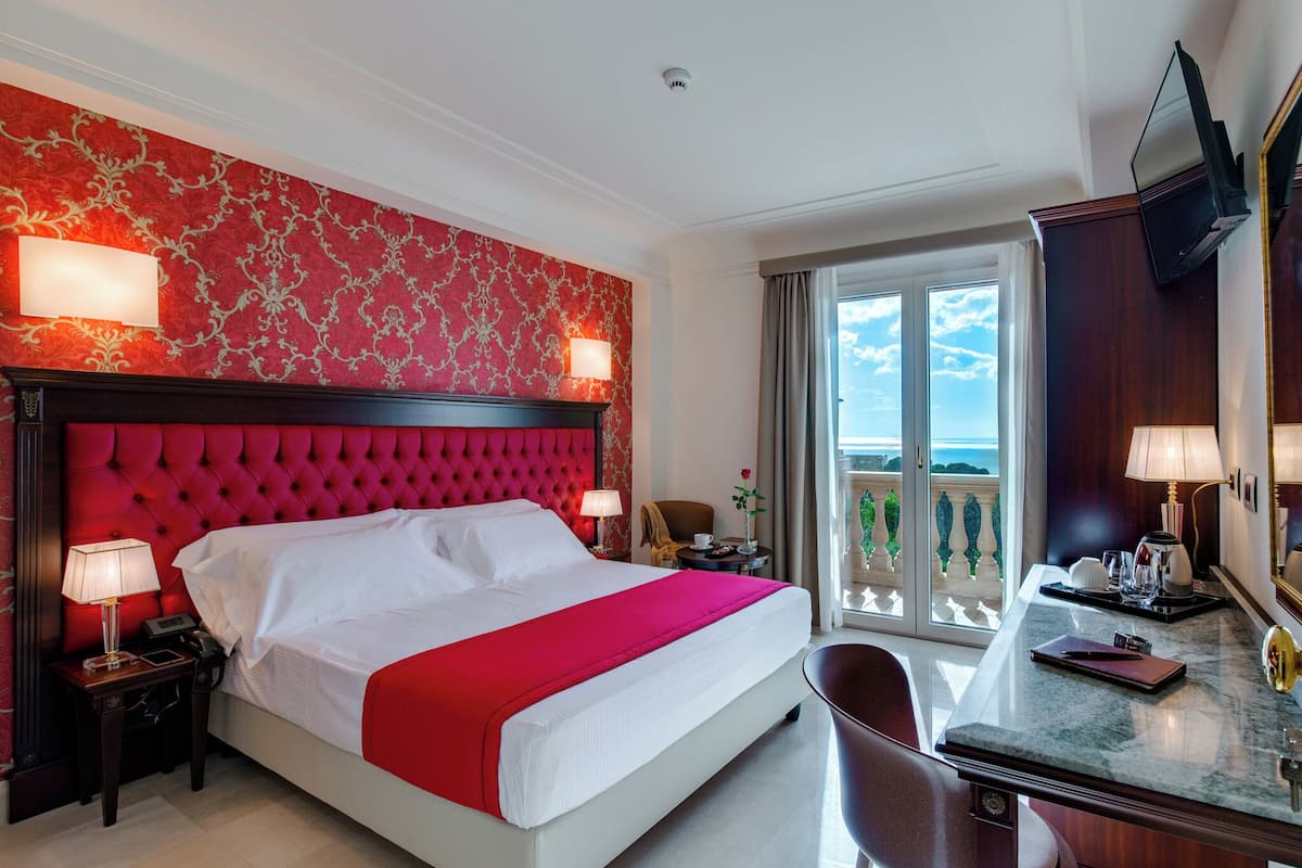 A hotel room with a large bed, a desk with a chair, a balcony with a view, and a red patterned wall.