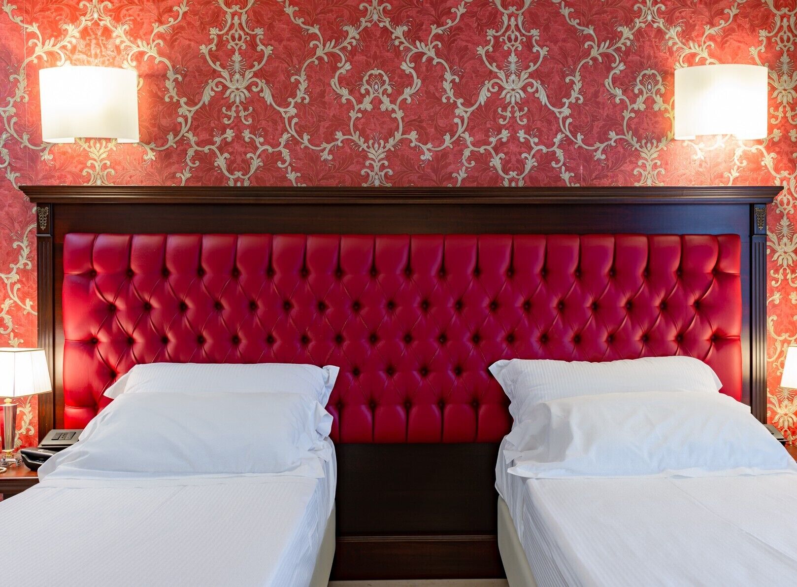 Una habitación de hotel con cabecera tapizada roja, dos camas blancas y papel tapiz decorativo.