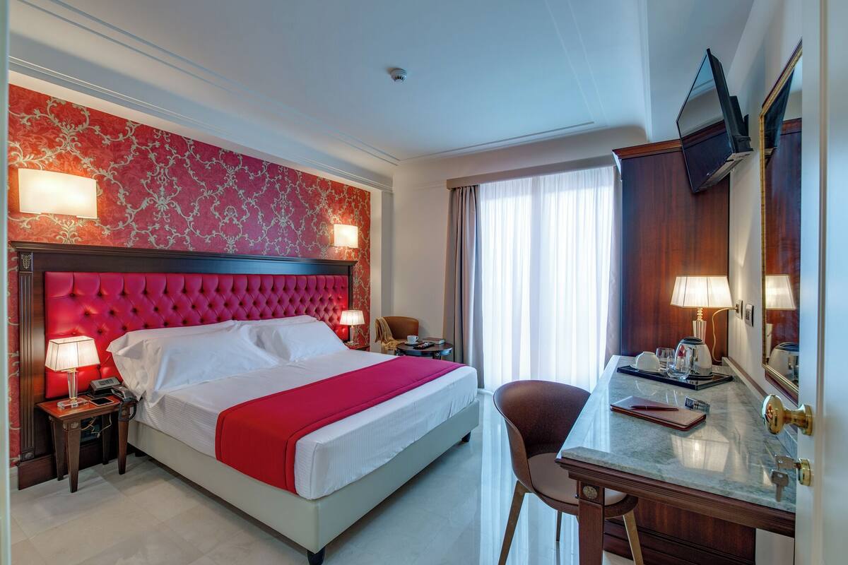 A hotel room with a large bed, a desk with a chair, a mirror, and a window with curtains.