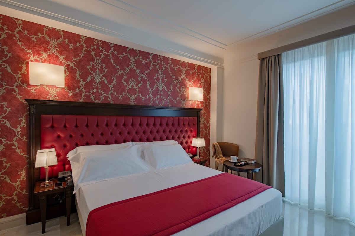 A hotel room with a large bed, red and gold patterned wallpaper, two bedside lamps, a red headboard, a small table with a lamp, and a view of a window with curtains.