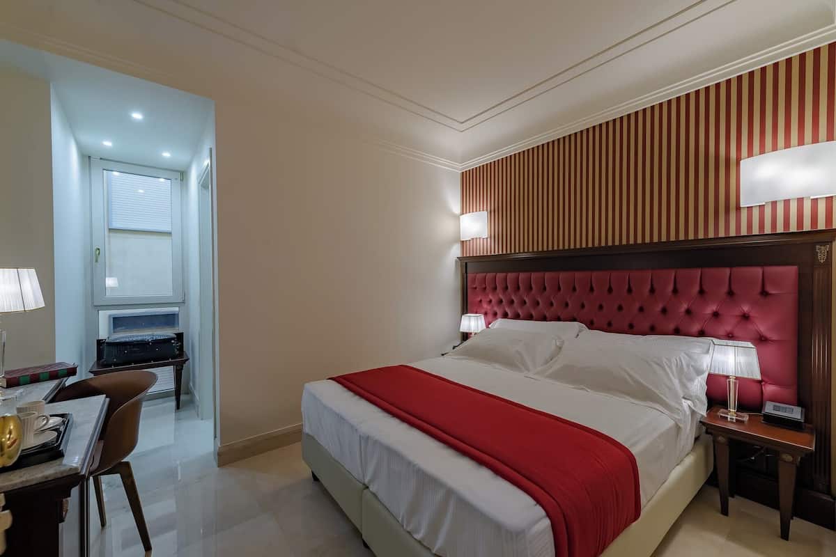 A hotel room with a large bed, a desk with a chair, and a small table with a lamp.