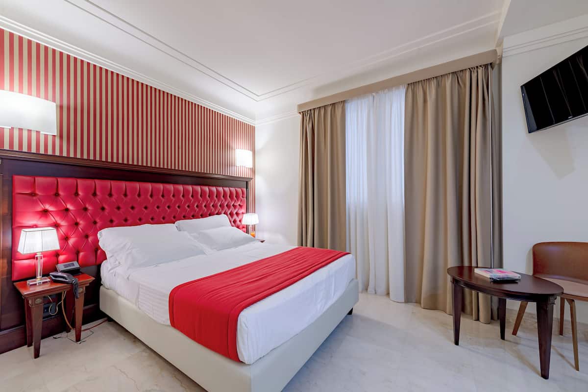 A hotel room with a large bed, a red headboard, a bedside table with a lamp, a TV mounted on the wall, and a window with curtains.