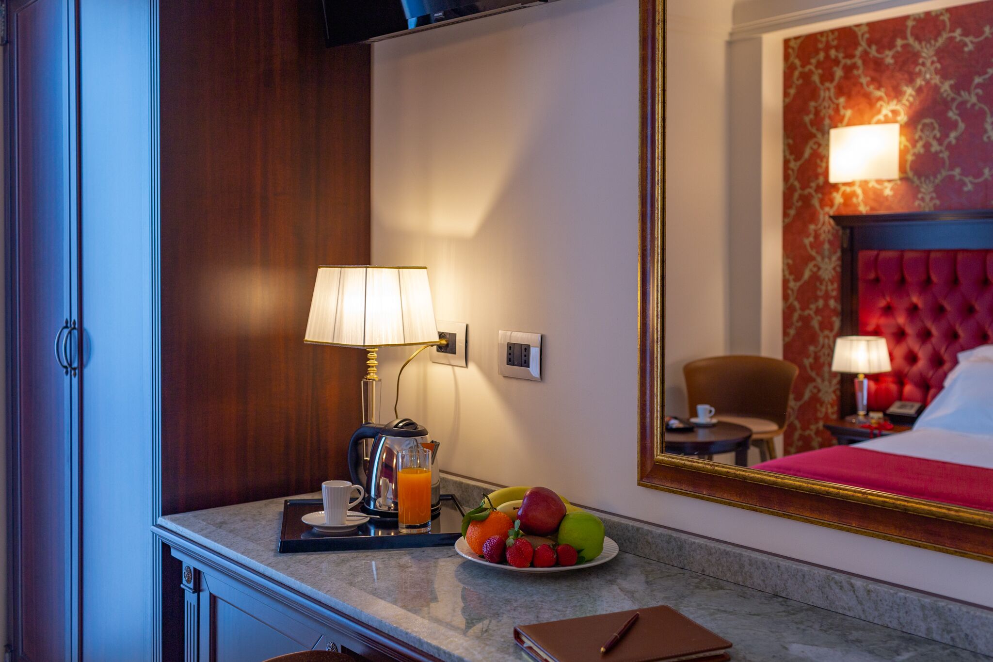 Une chambre d’hôtel avec un lit, une table de chevet avec une lampe, une assiette de fruits, une machine à café et un plateau avec une tasse et un verre de jus d’orange.