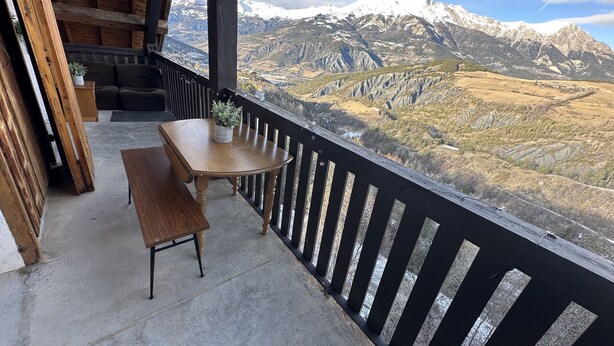 A wooden balcony with a table and bench, overlooking a mountainous landscape with snow-capped peaks. - Apartment in Le Sauze with unobstructed mountain views (Enchastrayes)