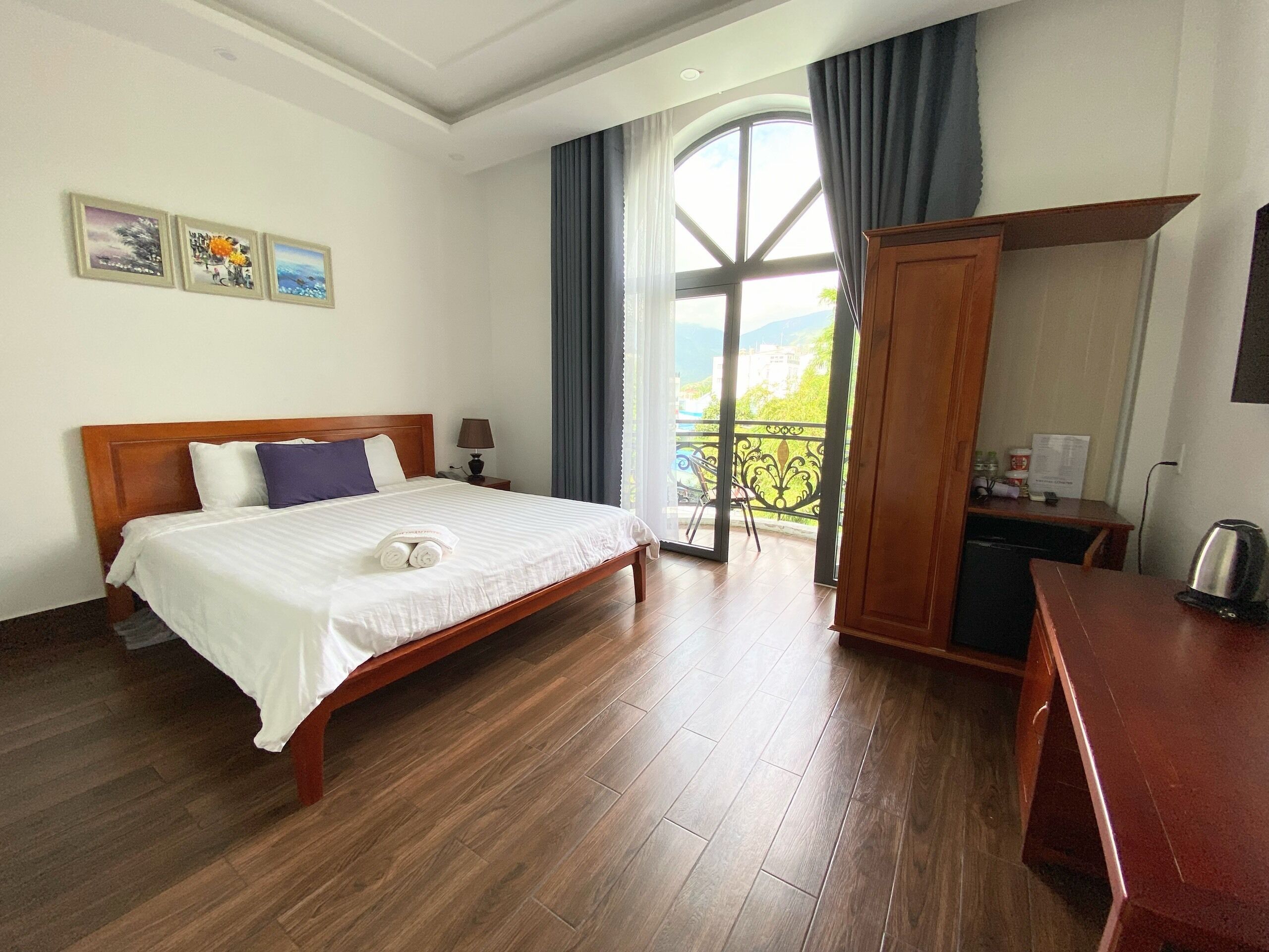 A hotel room with a wooden bed, a desk, a chair, a window with curtains, and a balcony with a view.