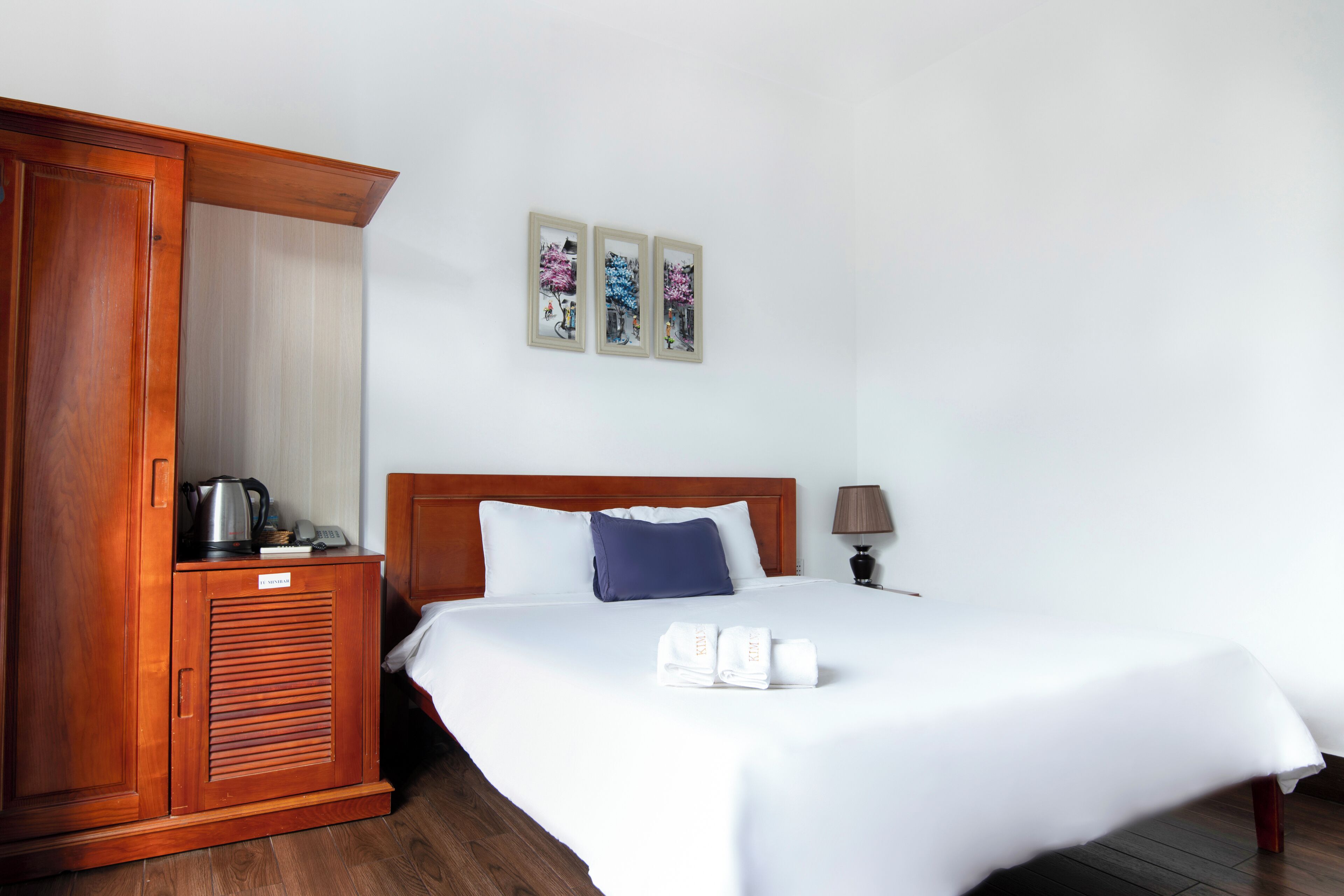 Une chambre d’hôtel avec une armoire en bois, un lit avec du linge de maison blanc, un oreiller bleu et deux tableaux encadrés au mur.