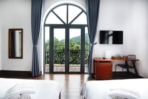 Habitación de hotel con dos camas, un escritorio, una silla, un televisor y un balcón con vistas a zonas verdes.
